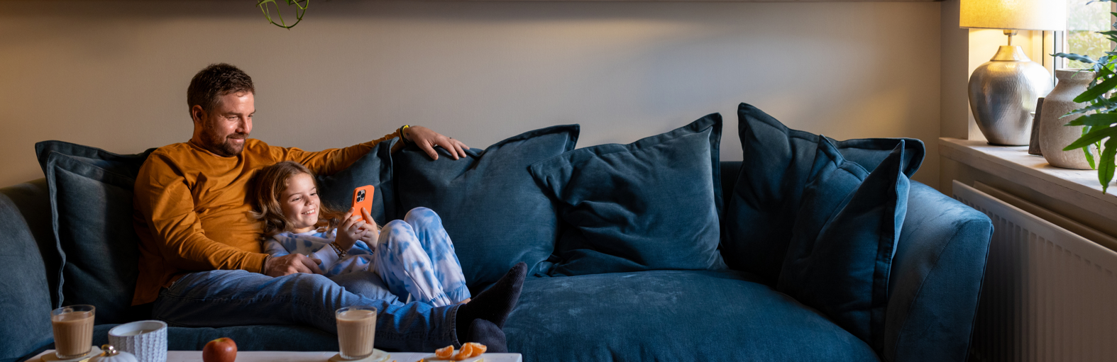 A full shot of a father lying down on the sofa with his daughter lying next to him. The girl is holding a mobile phone with her father looking over her shoulder.