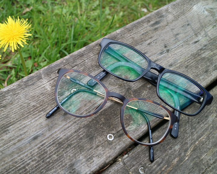 Två par glasögon ligger på en träplanka med grönskande gräs och maskros i bakgrunden