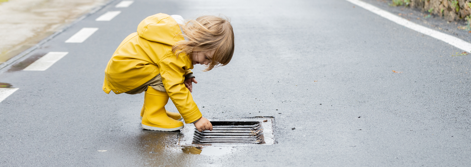 Side view of curious little child in vivid yellow raincoat and rubber boots standing on wet asphalt road after rain. Warm clothing concept for winter and rainy days